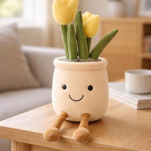 May include: A plush, smiling flower pot with yellow tulip flowers and green stems and leaves. The pot is beige with black eyes and a curved mouth. It has brown legs and sits on a wooden table.