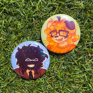 May include: Two circular pin-back buttons featuring stylized character illustrations. One button displays an orange-haired character with glasses, while the other shows a dark-haired character with a light-colored smile on a blue background.
