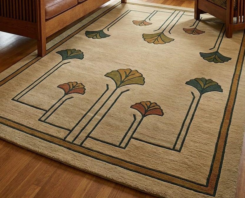 May include: A rectangular area rug with a tan background featuring a repeating pattern of stylized ginkgo leaf motifs in shades of green, orange, and brown. The design includes dark green linear borders and stems connecting the floral elements.