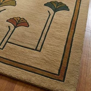 May include: A beige area rug featuring a repeating pattern of stylized fan-shaped floral motifs in shades of green, orange, and brown. The rug includes a dark green and orange border frame that outlines the central design with clean, straight lines.