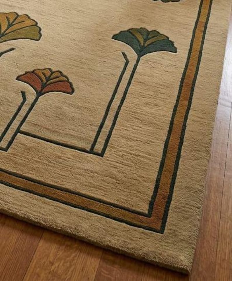 May include: A beige area rug featuring a repeating pattern of stylized fan-shaped floral motifs in shades of green, orange, and brown. The rug includes a dark green and orange border frame that outlines the central design with clean, straight lines.