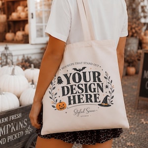 May include: A cream-colored canvas tote bag featuring a black graphic print that reads YOUR DESIGN HERE, surrounded by illustrations of a bat, a ghost, a spider, a pumpkin, and a witch hat, with the text MOCKUPS THAT STAND OUT above the design.