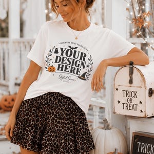 May include: A white short-sleeve t-shirt featuring a black graphic print that reads "YOUR DESIGN HERE" surrounded by illustrations of a pumpkin, a witch hat, a ghost, a bat, and foliage. The shirt is paired with a brown and black leopard print skirt.