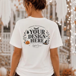 May include: A white t-shirt featuring a circular Halloween-themed graphic on the back. The design includes the text "YOUR DESIGN HERE" in black, surrounded by illustrations of a bat, a ghost, a spider, a pumpkin, and a witch hat with foliage.