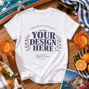 Könnte beinhalten: Ein weißes kurzärmeliges T-Shirt mit dem dunkelblauen Text "MOCKUPS THAT STAND OUT YOUR DESIGN HERE" und "Styled Scene STUDIO", umgeben von einer Blattgrafik. Das T-Shirt liegt auf einem Holzdeck mit Orangen und einer Sonnenbrille.