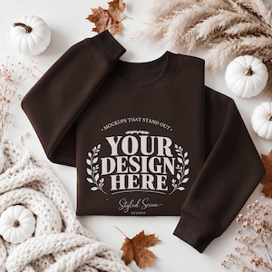 May include: A dark brown crewneck sweatshirt featuring the white printed text "MOCKUPS THAT STAND OUT" at the top, "YOUR DESIGN HERE" in the center, and "Styled Scene STUDIO" at the bottom, framed by two symmetrical white leaf branches.