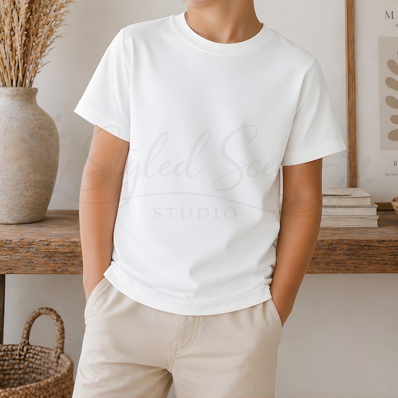 Puede incluir: Una camiseta blanca de cuello redondo y pantalones cortos color caqui. La camiseta es lisa y tiene mangas cortas. La persona est&aacute; de pie con las manos en los bolsillos. El fondo incluye un jarr&oacute;n, una cesta y un estampado enmarcado.