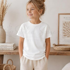 Puede incluir: Una persona joven con una camiseta blanca lisa de cuello redondo y pantalones color beige. La persona est&aacute; de pie frente a una estanter&iacute;a de madera con libros, un jarr&oacute;n con plantas secas y un grabado enmarcado. La camiseta tiene mangas cortas.