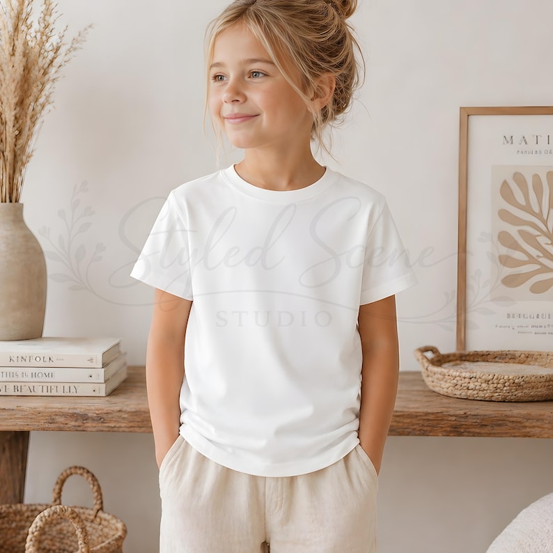 Puede incluir: Una persona joven con una camiseta blanca lisa de cuello redondo y pantalones color beige. La persona est&aacute; de pie frente a una estanter&iacute;a de madera con libros, un jarr&oacute;n con plantas secas y un grabado enmarcado. La camiseta tiene mangas cortas.