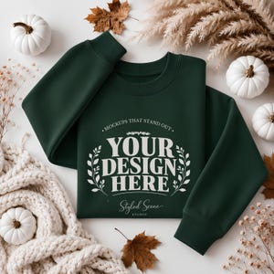 Könnte beinhalten: Ein dunkelwaldgrüner Rundhals-Pullover mit weißem Siebdruck-Text, der "MOCKUPS THAT STAND OUT" über "YOUR DESIGN HERE" und "Styled Scene STUDIO" am unteren Rand liest, eingerahmt von zwei dekorativen weißen Illustrationen von Blattzweigen.