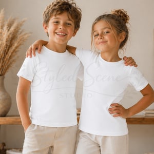 Puede incluir: Dos ni&ntilde;os con camisetas blancas y pantalones beige posan para una foto. El ni&ntilde;o tiene el pelo casta&ntilde;o y la ni&ntilde;a tiene el pelo recogido en un mo&ntilde;o. Las camisetas son de cuello redondo y manga corta. La imagen es luminosa.