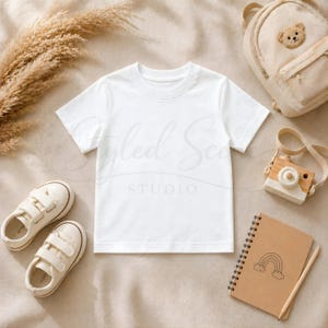 Puede incluir: Una camiseta blanca, zapatos de ni&ntilde;o, una mochila con dise&ntilde;o de oso, una c&aacute;mara de juguete de madera y un cuaderno con una ilustraci&oacute;n de arco&iacute;ris est&aacute;n dispuestos sobre una superficie beige. La imagen tiene una paleta de colores suave y neutra, adecuada para productos infantiles.