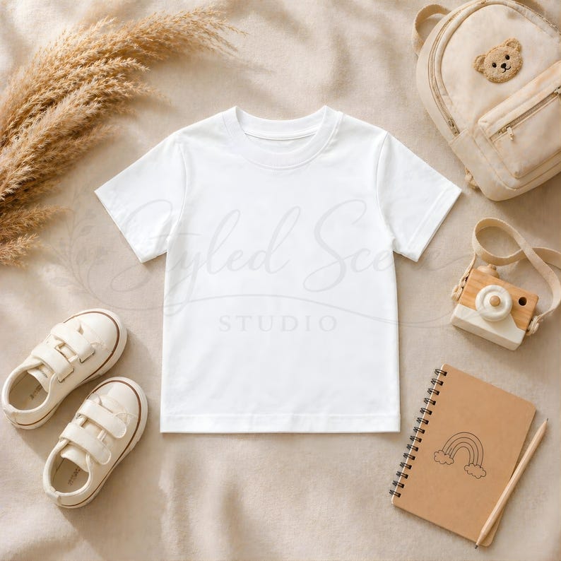 Puede incluir: Una camiseta blanca, zapatos de ni&ntilde;o, una mochila con dise&ntilde;o de oso, una c&aacute;mara de juguete de madera y un cuaderno con una ilustraci&oacute;n de arco&iacute;ris est&aacute;n dispuestos sobre una superficie beige. La imagen tiene una paleta de colores suave y neutra, adecuada para productos infantiles.