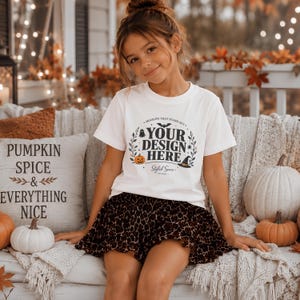 May include: A white short-sleeve t-shirt featuring a black graphic print that reads YOUR DESIGN HERE surrounded by illustrations of a pumpkin, a witch hat, a ghost, a bat, and foliage, paired with a brown and black leopard print skirt.