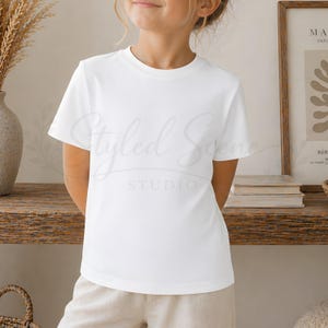 Puede incluir: Una camiseta blanca de cuello redondo. La camiseta de manga corta es lisa y de dise&ntilde;o sencillo. La modelo lleva pantalones beiges. El fondo incluye una estanter&iacute;a de madera con libros y un estampado enmarcado.