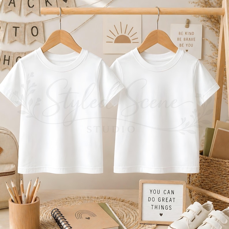 Puede incluir: Dos camisetas blancas colgadas de perchas de madera. Las camisetas se exhiben sobre un fondo con art&iacute;culos de papeler&iacute;a, una mochila y un letrero que dice "YOU CAN DO GREAT THINGS". La imagen tiene una est&eacute;tica limpia y minimalista.
