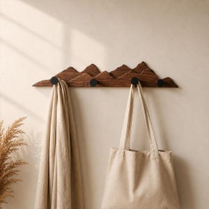 Könnte beinhalten: Ein Wandgarderobe aus Holz mit einem Bergketten-Design. Die Garderobe ist aus dunkelbraunem Holz gefertigt und verfügt über vier schwarze Haken. Eine beige Tragetasche und ein beiger Schal hängen an den Haken.