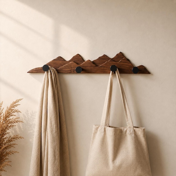 May include: A wooden wall-mounted coat rack with a mountain range design. The rack is made of dark brown wood and features four black hooks. A beige tote bag and a beige scarf hang from the hooks.