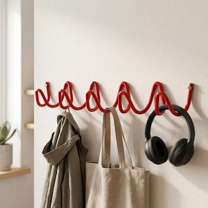 Può includere: Un attaccapanni da parete in metallo rosso dal design ondulato unico. Una giacca grigia, una borsa tote beige e delle cuffie nere sono appese ai ganci. L'attaccapanni è montato su una parete bianca, offrendo una soluzione di archiviazione moderna.