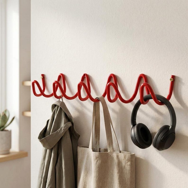 May include: A red metal wall-mounted coat rack with a unique, wavy design. A gray jacket, a beige tote bag, and black headphones hang from the hooks. The rack is mounted on a white wall, offering a modern storage solution.