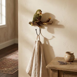 May include: A decorative wall hook featuring a bird perched on a branch. The bird is green, brown, and yellow. A black hook hangs below the branch, holding a beige towel. The hook is mounted on a light-colored wall.