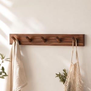 May include: A dark brown wooden coat rack with five pegs, mounted on a wall. A beige towel and a beige net bag are hanging from the pegs. The rack is rectangular. The background is a plain white wall. The rack is suitable for a hallway or bathroom.