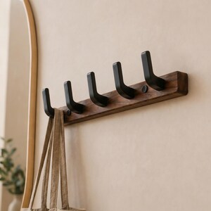 May include: A wooden wall-mounted coat rack with five black hooks. The rack is made of dark brown wood and is mounted on a beige wall. A beige tote bag hangs from one of the hooks. The hooks are evenly spaced.