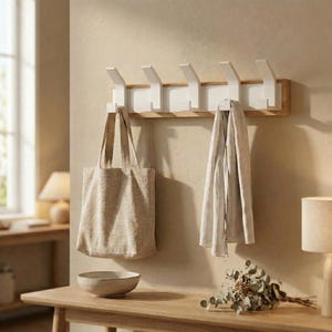 Op de afbeelding: Een houten wandkapstok met zes witte haken. Een beige draagtas en een lichtgekleurde sjaal hangen aan de haken. Een kleine, lichtgekleurde kom staat op een houten tafel.