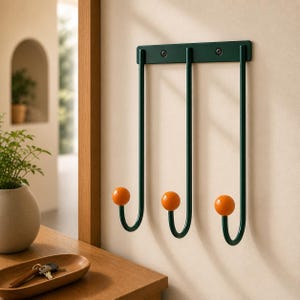 May include: A dark green metal wall hook with three curved arms, each ending in an orange sphere. The hook is mounted on a light-coloured wall. The design is modern and minimalist, suitable for a hallway or entryway.