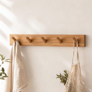 May include: A wooden wall-mounted coat rack with five pegs. A beige towel hangs from one peg, and a beige string bag hangs from another. The rack is made of light-colored wood and is mounted on a white wall.