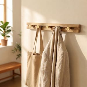 May include: A wooden coat rack with five brass hooks, mounted on a wall. A beige tote bag and a light-coloured linen jacket hang from the hooks. The rack is fixed to a neutral-toned wall, with natural light in the background.