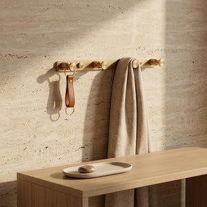 May include: A wooden coat rack with six brass hooks. A brown leather keyring hangs from one hook, and a beige towel is draped over another. A small, oval tray with a stone sits on a wooden bench below.