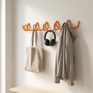 May include: An orange metal wall-mounted coat rack with a wavy design. A beige tote bag, black headphones, and a light gray jacket hang from the rack. The rack is mounted on a white wall.