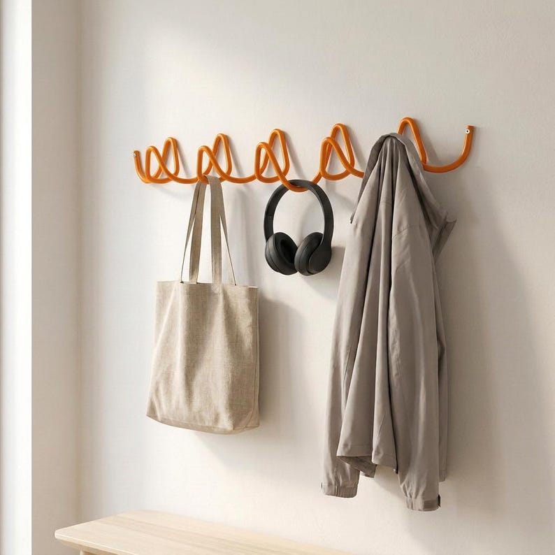 May include: An orange metal wall-mounted coat rack with a wavy design. A beige tote bag, black headphones, and a light gray jacket hang from the rack. The rack is mounted on a white wall.