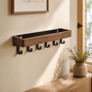 May include: A wall-mounted shelf with six black hooks. The shelf has a dark wood finish with a black interior and is mounted on a beige wall. A framed picture hangs above the shelf.