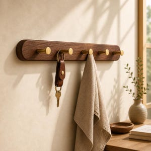 May include: A wooden wall-mounted key and towel rack with six brass hooks. A brown leather key holder with a key hangs from one hook, and a beige towel hangs from another. The rack is mounted on a light-colored wall.