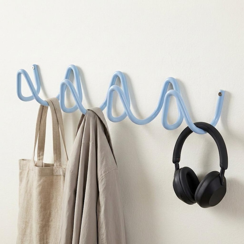 May include: A light blue metal wall-mounted coat rack with a wavy design. A beige tote bag, a light gray jacket, and black headphones hang from the rack. The rack is mounted on a white wall.