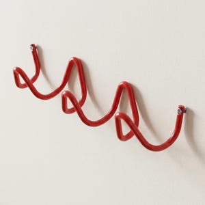 May include: A red metal wall-mounted coat rack with a wavy design. The rack is attached to a white wall with screws. The design features three curved sections, providing multiple hanging points for coats, hats, or other items.