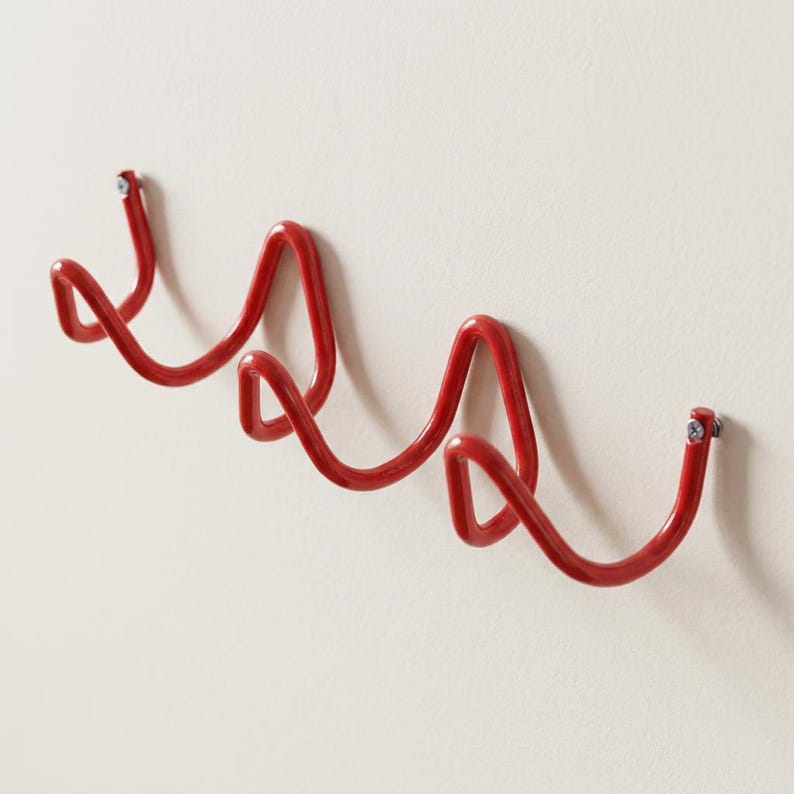 May include: A red metal wall-mounted coat rack with a wavy design. The rack is attached to a white wall with screws. The design features three curved sections, providing multiple hanging points for coats, hats, or other items.