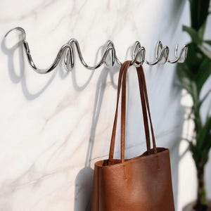 May include: A polished chrome wall-mounted coat rack with a unique, flowing design. A brown leather tote bag hangs from one of the hooks. The rack is mounted on a white marble-patterned wall, with a green plant visible in the background.