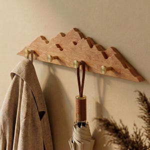 Puede incluir: Perchero de pared de madera con forma de montaña y ganchos de latón. El perchero es de madera marrón claro y tiene un diseño de cordillera. Un paraguas con un asa de cuero marrón y un abrigo beige cuelgan de los ganchos.