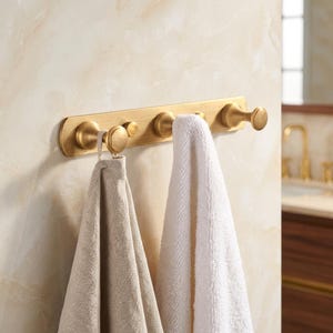 Può includere: Un portasciugamani in metallo color oro con cinque ganci arrotondati. Due asciugamani, uno beige e uno bianco, sono appesi ai ganci. Il portasciugamani è montato su una parete chiara e strutturata, suggerendo un ambiente bagno.
