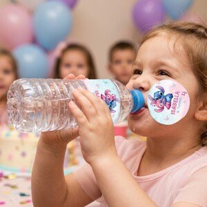 A young girl with brown hair drinks from a plastic water bottle at a birthday party, surrounded by blurred pastel balloons and children. The bottle features a custom pink and blue birthday label with Stitch and Angel, matching a round sticker on her cheek that reads "Isabella is turning 4."