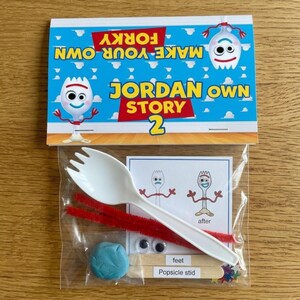 A flat-lay photograph on a wooden table shows a plastic bag attached to a printed paper card. The card features cartoon figures and text against a blue sky background. The text reads, "MAKE YOUR OWN FORKY," "JORDAN OWN STORY," and "2." The transparent plastic bag contains various craft supplies, including a white plastic spoon, red pipe cleaners, colorful popsicle sticks, googly eyes, a small ball of blue modeling dough, and a small instructional card.