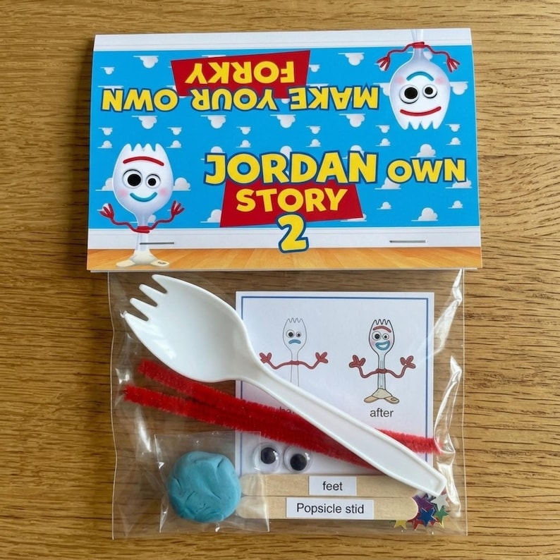 A flat-lay photograph on a wooden table shows a plastic bag attached to a printed paper card. The card features cartoon figures and text against a blue sky background. The text reads, "MAKE YOUR OWN FORKY," "JORDAN OWN STORY," and "2." The transparent plastic bag contains various craft supplies, including a white plastic spoon, red pipe cleaners, colorful popsicle sticks, googly eyes, a small ball of blue modeling dough, and a small instructional card.