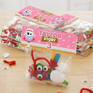 A close-up shot of assembled party favor bags stacked on a light wooden table. The front bag features a pink topper with a cloud pattern, an illustration of the Forky character, and the text "Taylor Story 3." Inside the clear plastic bag are visible DIY craft supplies, including googly eyes, colorful sticks, red pipe cleaners, and a ball of white clay.