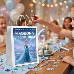A lifestyle mockup showing a white treat bag on a party table. The bag features a "Frozen" Elsa design for "Madison’s 6th Birthday." A child’s hand is placing a Woody from "Toy Story" action figure into the bag. The background is a festive party setting with blurred children, cake pops, and fairy lights.