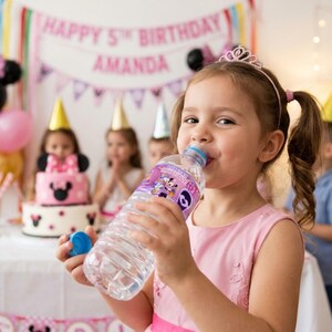 A young girl in a pink dress and a tiara happily drinks from a water bottle with a custom Minnie Mouse label. In the blurred background, other children sit at a party table with a Minnie Mouse-themed birthday cake and a banner that reads "Happy 5th Birthday Amanda." The setting is a bright, decorated indoor birthday party.