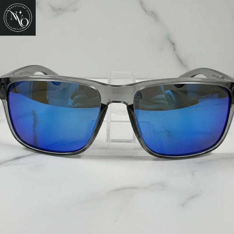 Pu&ograve; includere: Occhiali da sole grigi con lenti a specchio blu. Le montature sono di colore grigio traslucido. Le lenti riflettono una tonalit&agrave; blu brillante. Gli occhiali da sole sono esposti su un supporto trasparente su sfondo di marmo bianco.