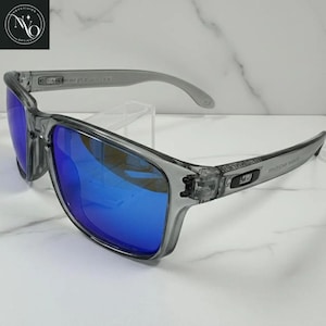 Pu&ograve; includere: Occhiali da sole grigi con lenti a specchio blu. Le montature sono grigie traslucide e le lenti riflettono una tonalit&agrave; blu brillante. Le aste hanno testo e un piccolo logo. Gli occhiali da sole sono esposti su una superficie bianca.